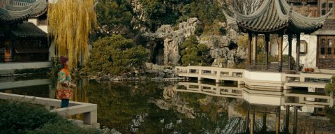 a woman holds a camera at a traditional Chinese garden with a pond, pavilion and trees