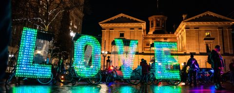 a night image of the word LOVE spelled out in large letters made of hundreds of small bulbs attached to bicycles in front of a large brick building