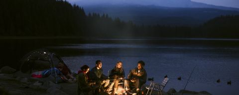 four people illuminated by campfire at a dimly lit campsite, backed by a serene lake and a large mountain obscured by mist