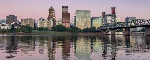 pale pink and purple sky reflect on a river surface with a bridge off to the side and a view of downtown in the background