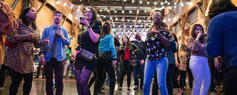 young people stand with drinks in their hands, string lights above in chic venue