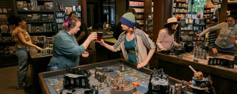 Two friends clinking glasses over a large tabletop board game.