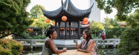 Two people sit at a table drinking tea with a Chinese pavilion in the background.