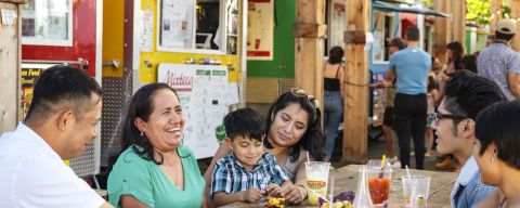 people sitting at a picnic table in front of a line of colorful food carts