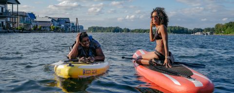 two women on paddleboards in a river with houseboats in the distance