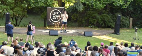Blond comedian on stage performing in front of large crowd sitting in a park