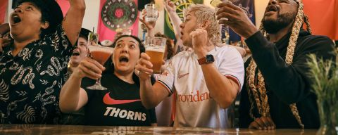 a crowd of people cheer while raising glasses in front of a bar while watching a game