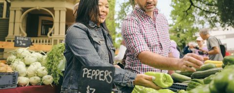 two people peruse fresh produce like peppers and zucchini outside in bins with signs saying what each bin contains and how much they are a pound