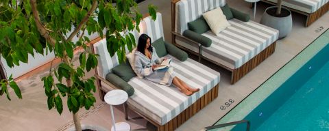 Woman in robe lounges next to a pool while reading a book.