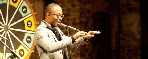 a man speaking on stage with a gameshow wheel behind him