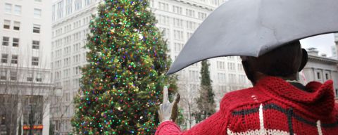 a large tree decorated with lights in front of a statue of a person holding an umbrella