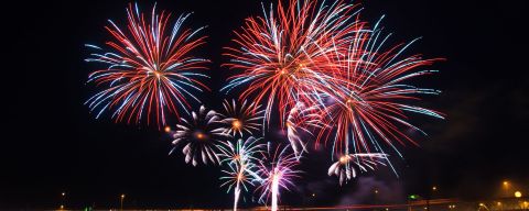 Fireworks burst in a night sky above a river, with boats in the foreground and a bridge in the background