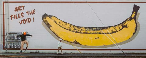 A man and a child skateboard on a sidewalk in front of a mural of a yellow banana with text reading. \"Art fills the void!\"