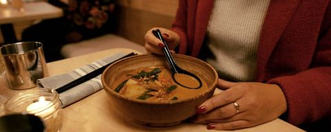 A person sitting at a table holds a ramen spoon in a bowl