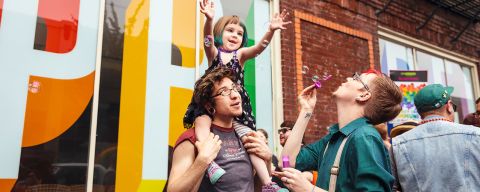 a parent carries their child on their shoulders while the other parent blows bubbles toward them