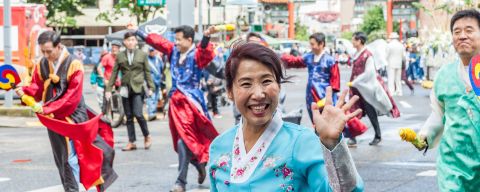 woman with big smile waves in a parade with colorful costumes on all the folks in the frame Chinese gate in background