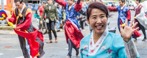 A woman wearing a hanbok and a big smile waves at the camera among a crowd of parade marchers in front of the ornate gate in Old Town China Town.