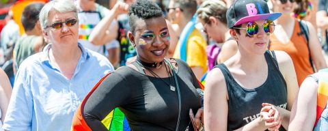 people stand watching a parade wearing rainbow flags and rainbow makeup with rainbows reflecting in their sunglasses
