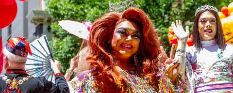 two drag queens smile and wave at the camera in front of yellow and red balloons