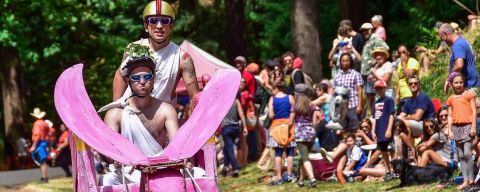 two people wearing sunglasses and togas ride a pink elephant-shaped soapbox derby cart