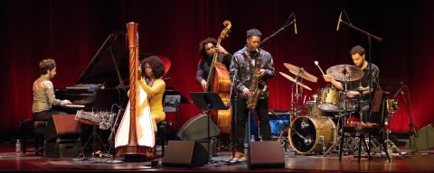 A band of five Jazz musicians performs on stage in front of a red curtain.