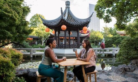 Two people sit at a table drinking tea with a Chinese pavilion in the background.