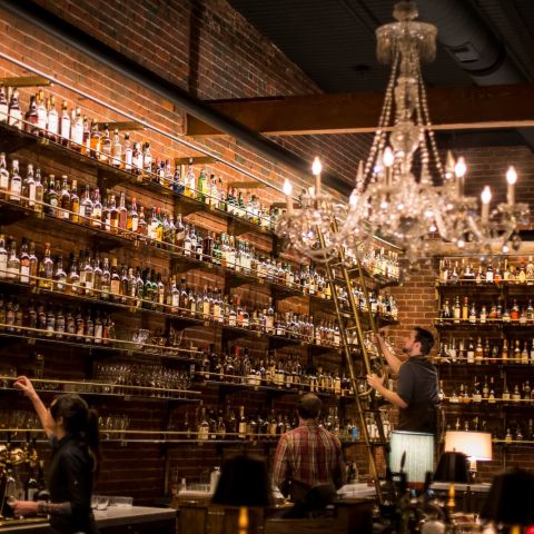 several bartenders peruse shelves housing a vast collection of whiskey bottles