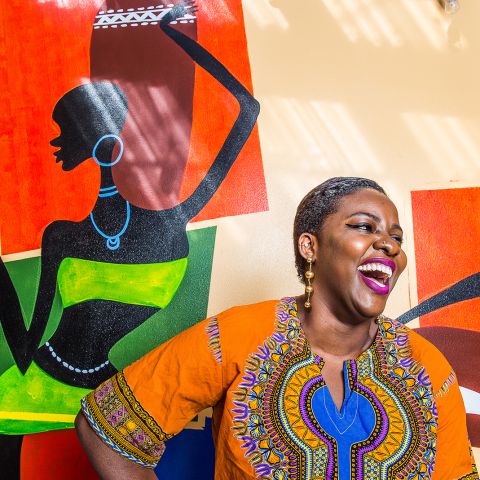 a Black woman wearing an orange dashiki stands in front of a colorful mural laughing