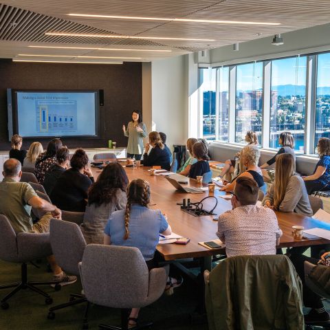 a large group of people around a conference room table watch a person giving a presentation
