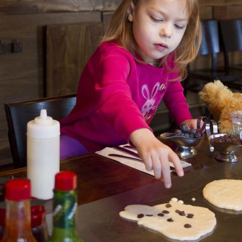 a young girl adds chocolate chips and blueberries to a pancake