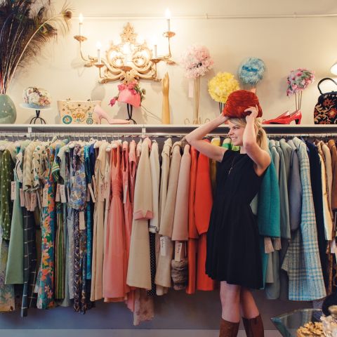 a woman trying on a hat in front of racks of vintage clothing