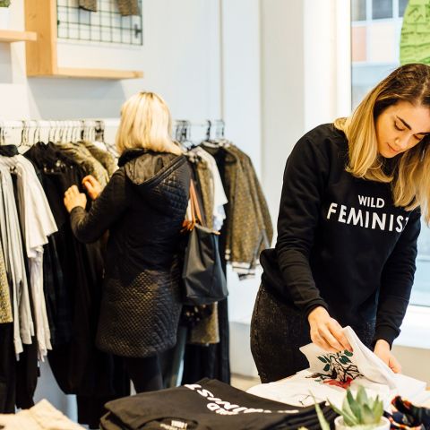 a person in a black shirt that reads “wild feminist” folds a shirt in a modern clothing store