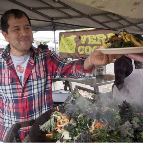 man in an outdoor market handing a plate of food to a customer in front of a sign reading \"Verde Cocina\"