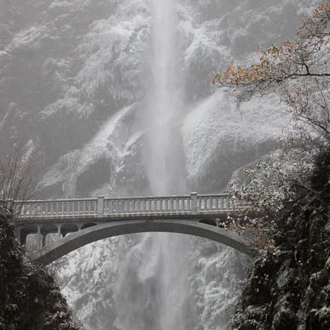 a towering waterfall behind a stone bridge surrounded by snow and ice