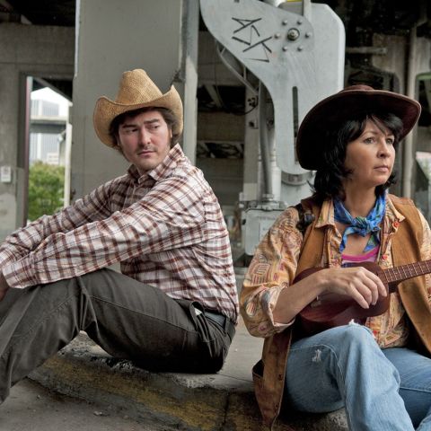 Two actors from the Fertile Ground Festival, dressed in cowboy hats, sit on a curb beneath a freeway overpass. She plays a small guitar as he looks on.