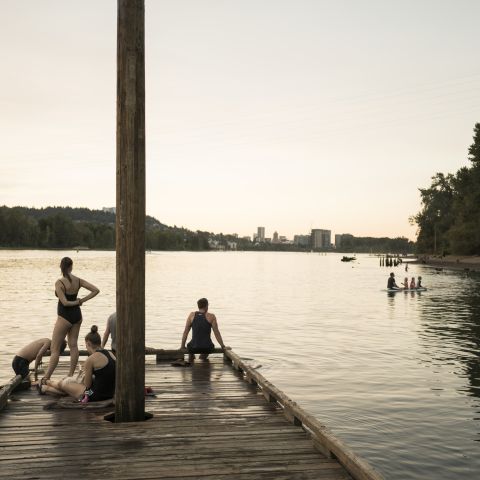 a wooden dock in a river — with swimmers standing, sitting or climbing out of the water — looks toward a city skyline in the fading sunlight