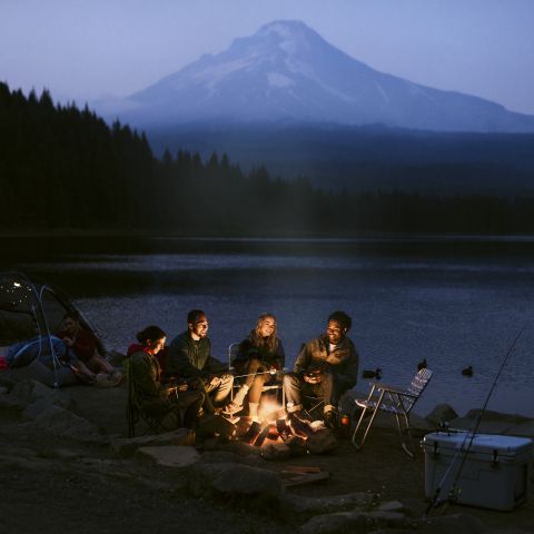 four people illuminated by campfire at a dimly lit campsite, backed by a serene lake and a large mountain obscured by mist