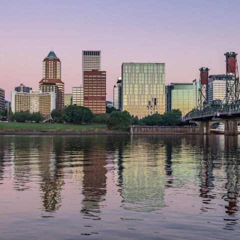 pale pink and purple sky reflect on a river surface with a bridge off to the side and a view of downtown in the background