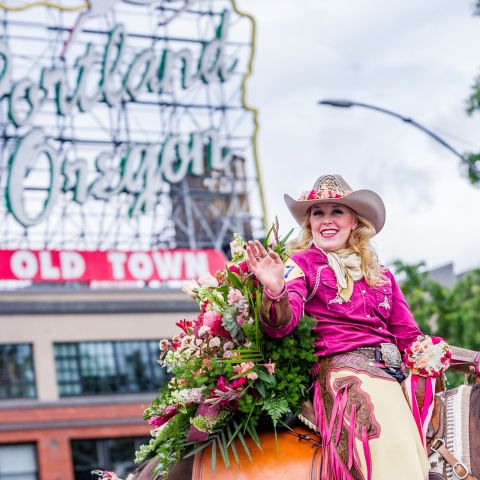 woman in hat and chaps ride a rose decorated horse in a parade with Portland\'s white stag sign in the background