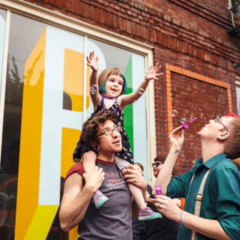 a parent carries their child on their shoulders while the other parent blows bubbles toward them