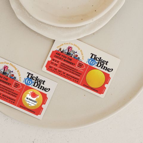 two colorful scratch-off tickets that read Ticket to Dine and Thank You For Dining With Us rest on a large white plate on a white countertop