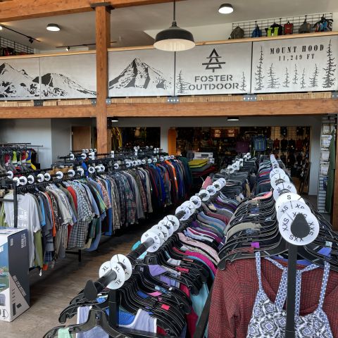 Racks of new and used outdoor clothing sit in a bright space accented by wood beams and the Foster Outdoor mountain scape logo.