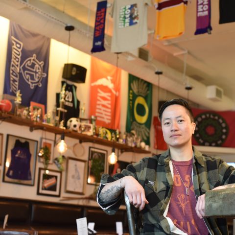 A woman in a plaid button-down and red t-shirt sits at a bar in front of a wall of women\'s sports memorabilia.