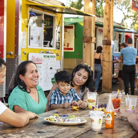 people sitting at a picnic table in front of a line of colorful food carts