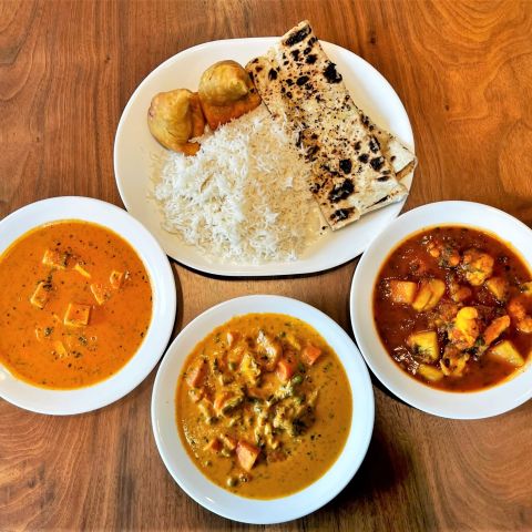 Three dishes of curries and aloo gobi sit next to a platter of rice, samosas and naan on a wood table.