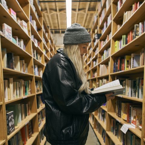 a person in a hat and leather jacket stands in-between towering stacks of books with a book in hand, reading