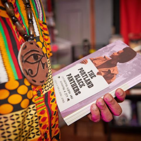 a close-up of a hand holding the book “The Portland Black Panthers” with bookshelves in the background