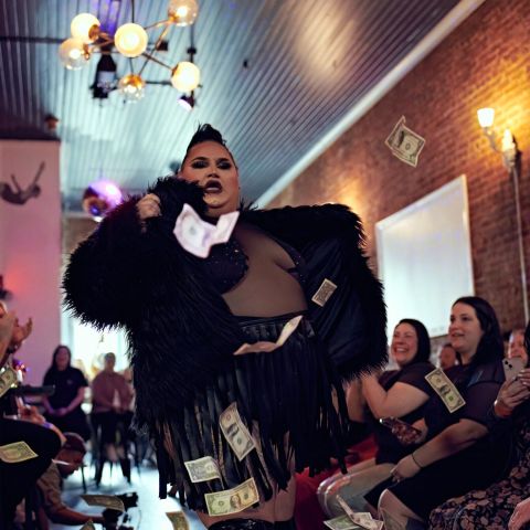 A woman in a sheer bedazzled body suit and black feather jacket performs amidst a crowd showering her with dollar bills.