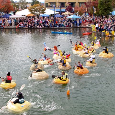 people in costumes paddle boats made of giant pumpkins on a lake while spectators watch on the shore
