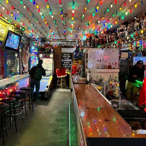 a patron plays Skee-Ball and a bartender leans against the backbar in a tavern lighted with Christmas lights